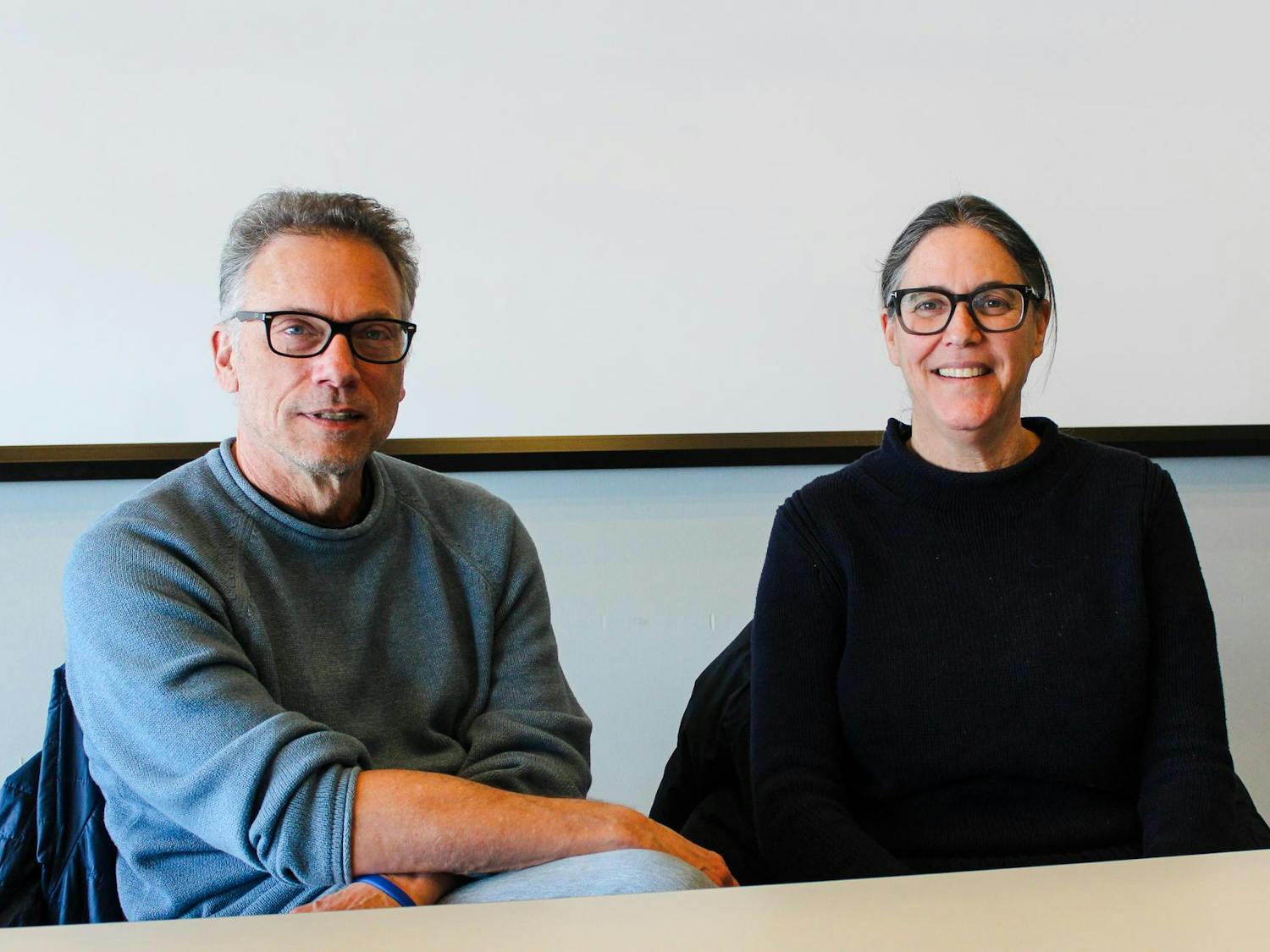 Visiting lecturers Jennifer Levin and John Eisendrath, wearing matching black-rimmed glasses and black and gray sweaters, smile against a white background.