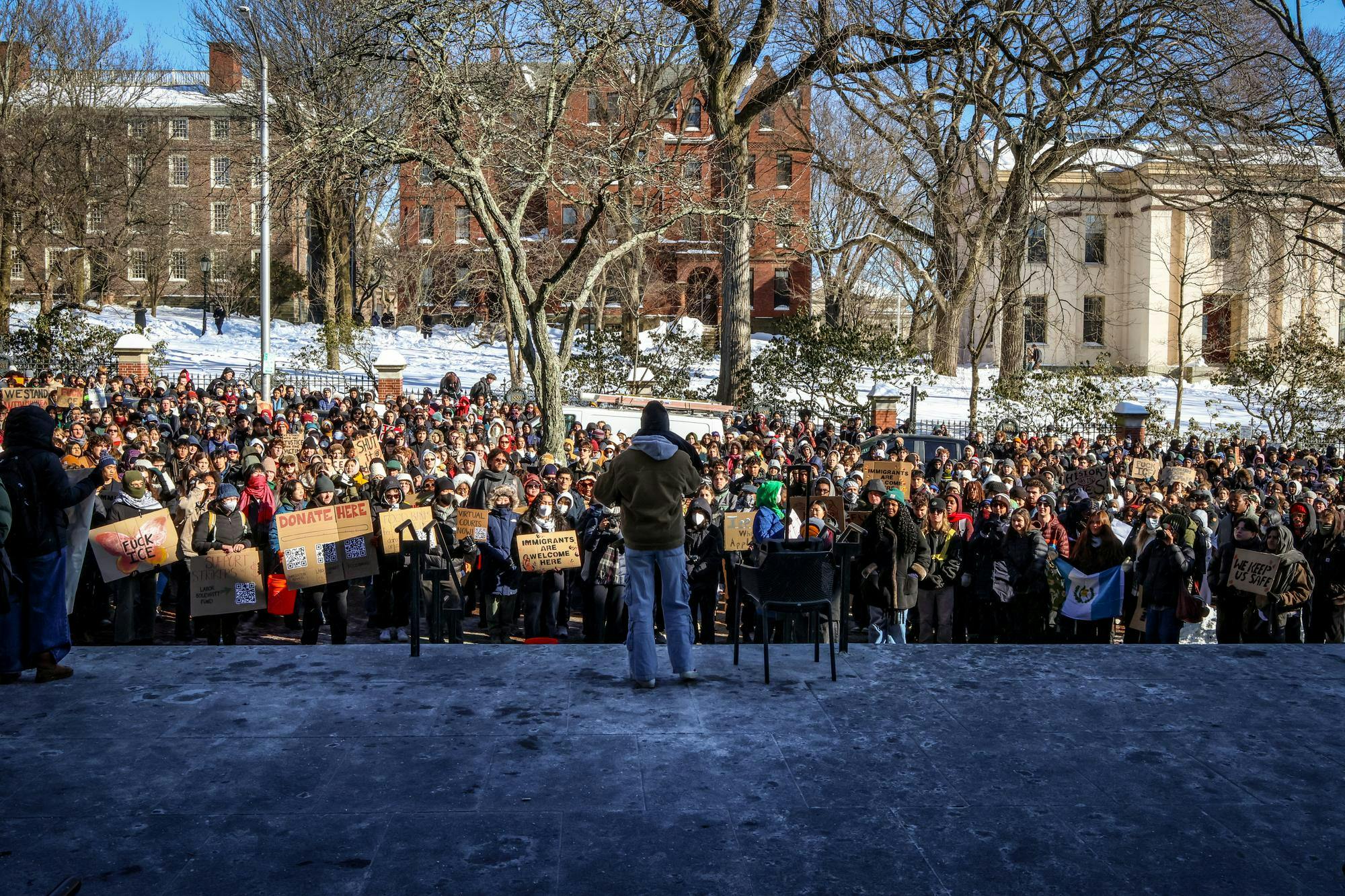 A man speaks to a large crowd on the steps of Brown University’s John D. Rockefeller Jr. Library. Many people within the crowd hold signs and wear masks.  