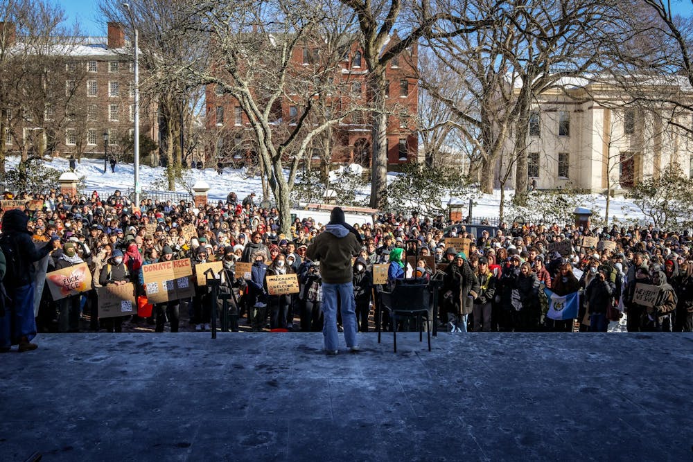 A man speaks to a large crowd on the steps of Brown University’s John D. Rockefeller Jr. Library. Many people within the crowd hold signs and wear masks.  