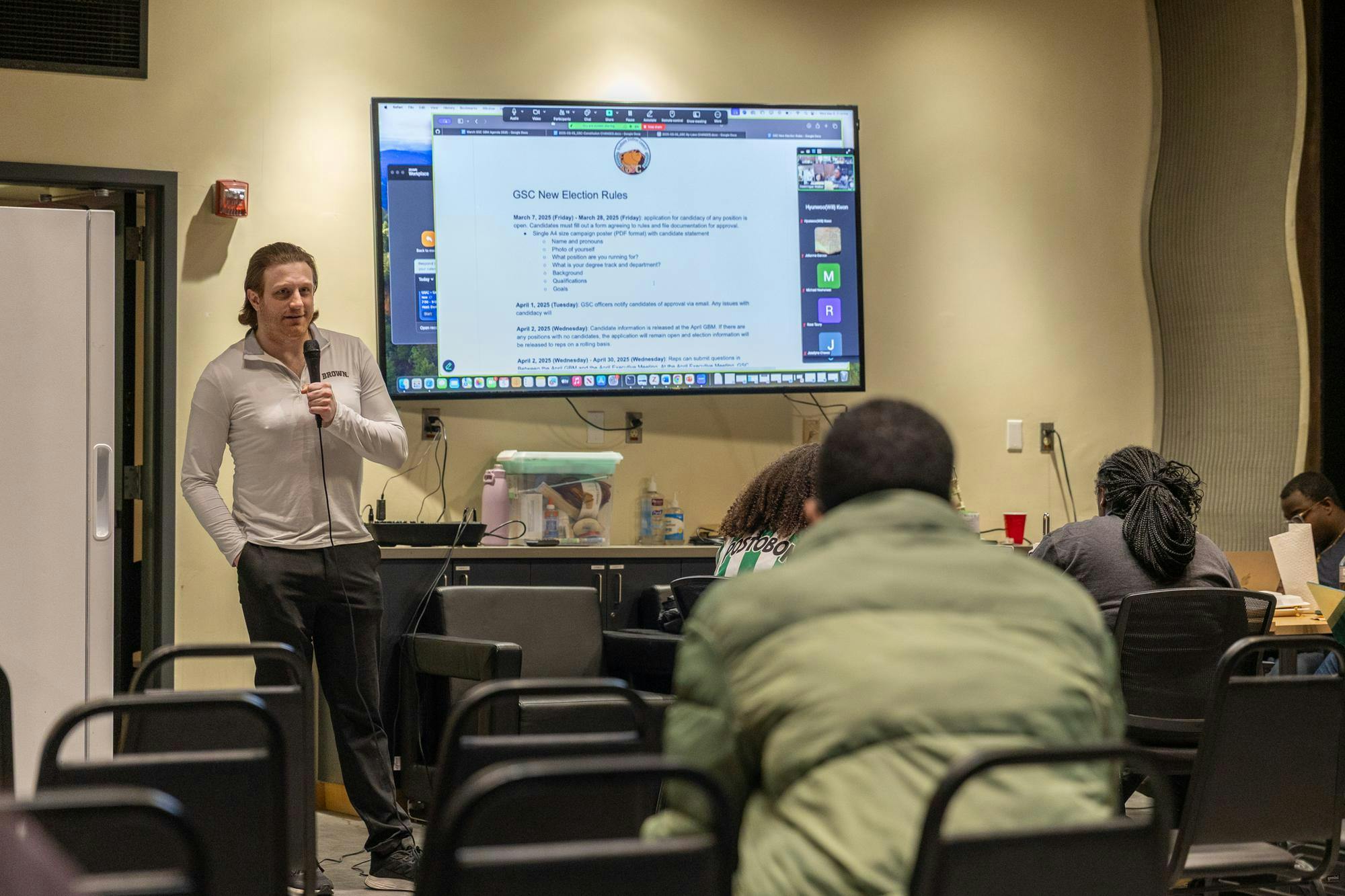 GSC President Kevin LoGiudice ScM’21 GS stands with a microphone in front of room full of graduate students presenting on a pull down screen. 
