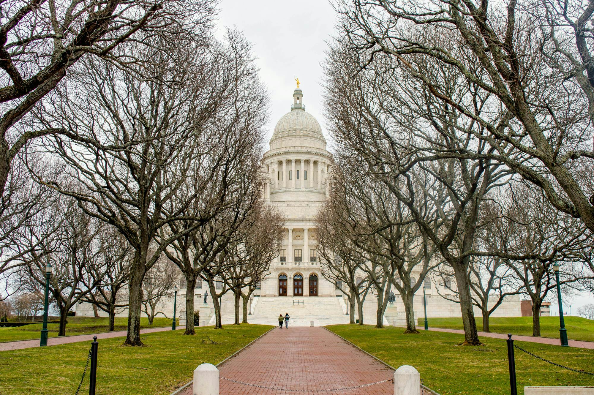 A photo shows the Rhode Island Statehouse from its entrance path, lined with leafless trees on a cloudy day. 

