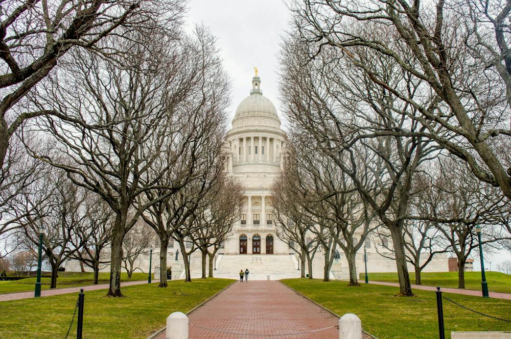A photo shows the Rhode Island Statehouse from its entrance path, lined with leafless trees on a cloudy day. 

