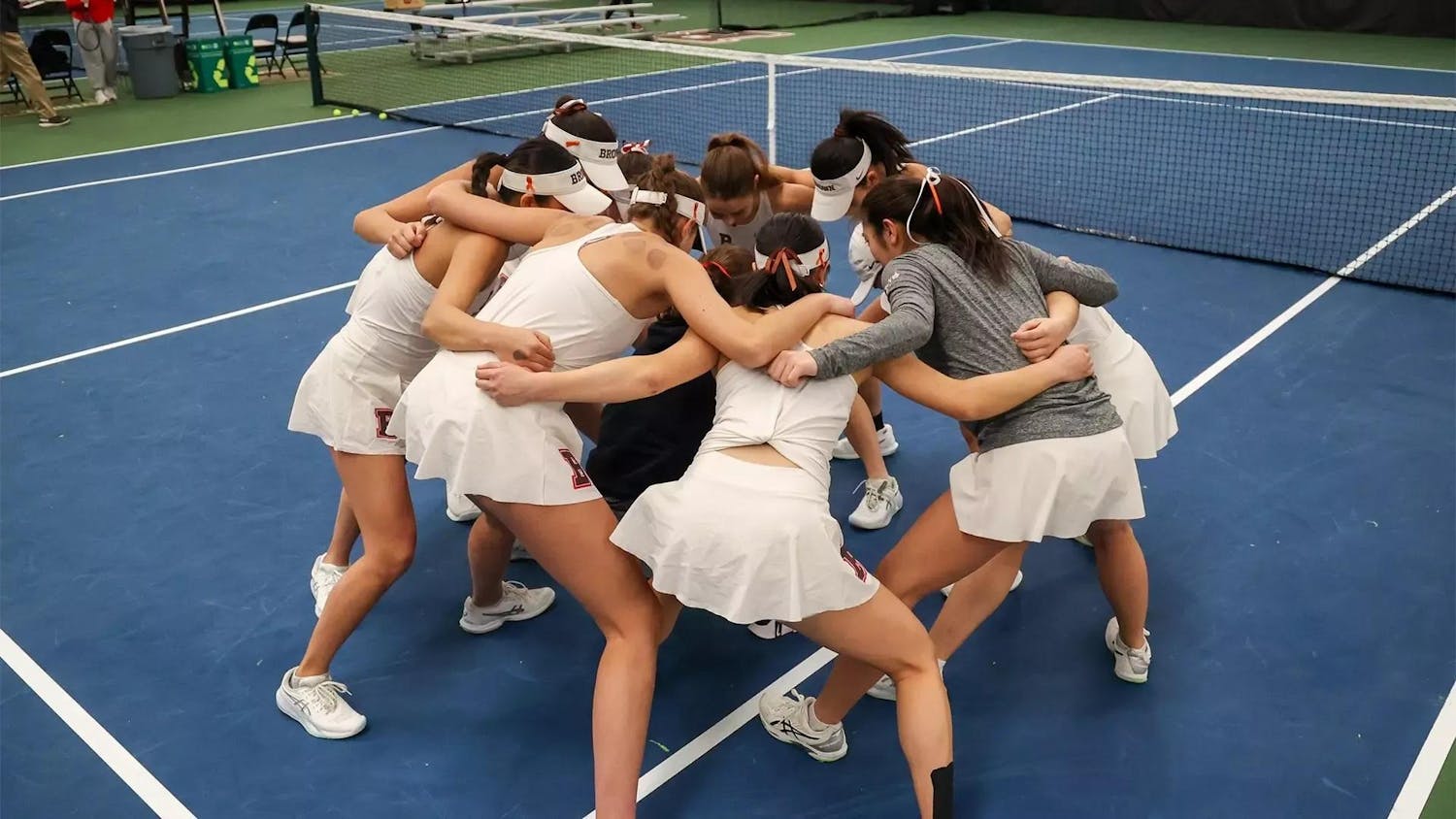 Photo of Brown women’s tennis players in white tank tops and skirts with the Brown University “B” on it, huddled in a circle with their arms around each other.