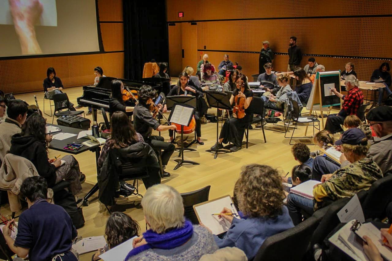 Audience members seated in an auditorium sketch on clipboards while student musicians play on the stage. 
