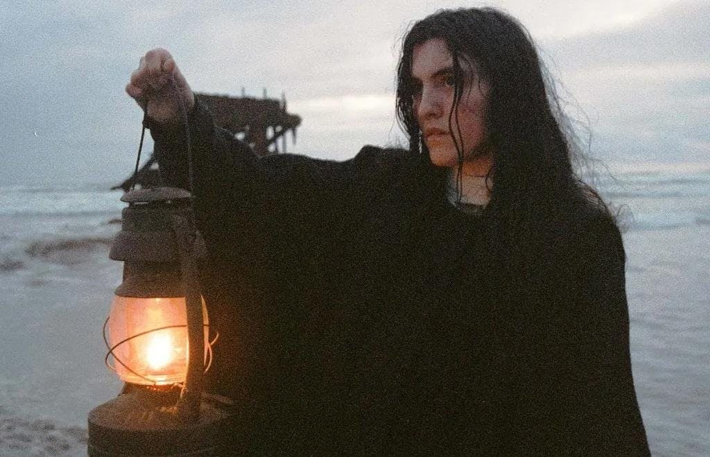 Photo of a person with long hair in a robe shining a lamp on a beach in gloomy weather.

