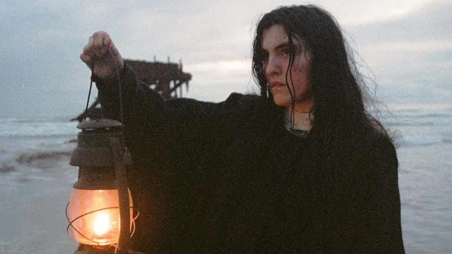 Photo of a person with long hair in a robe shining a lamp on a beach in gloomy weather.