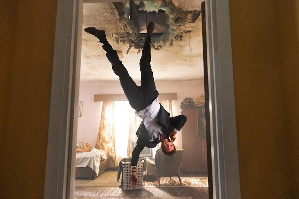 A picture of Jack Quaid in a suit upside down falling from a hole in the ceiling.