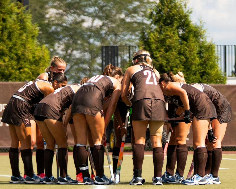 Photo of multiple Brown Field Hockey players huddled together on a field.