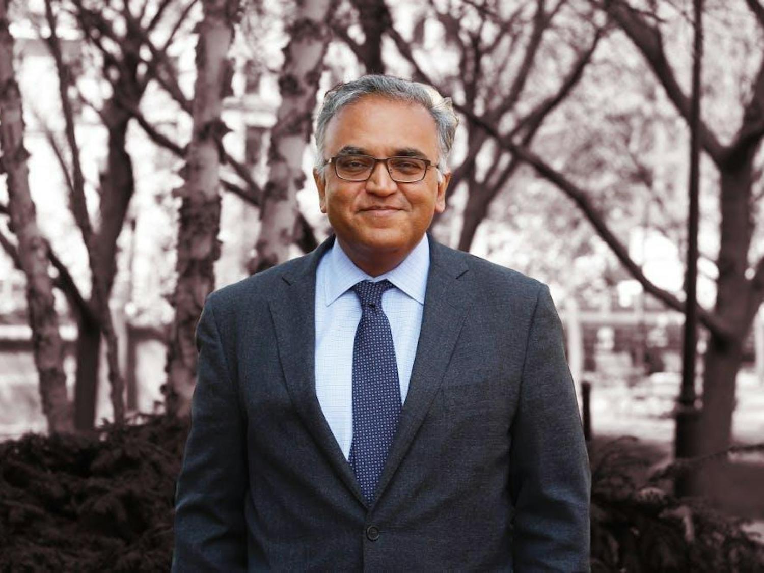A headshot of Ashish Jha wearing a suit with a blue tie in front of some trees.