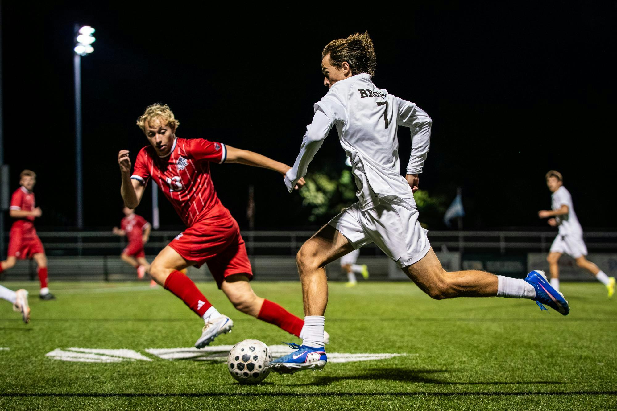 Photo showing Brown University soccer player in a white uniform running towards a ball with an NJIT player in a red uniform running alongside him on the field at night.  