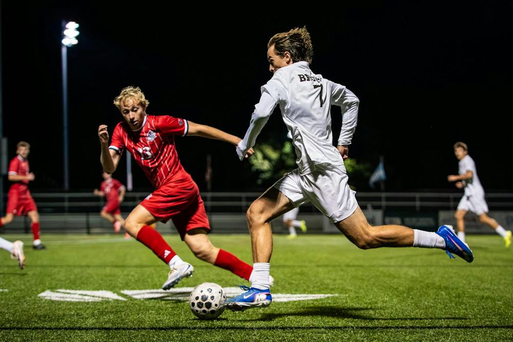 Photo showing Brown University soccer player in a white uniform running towards a ball with an NJIT player in a red uniform running alongside him on the field at night.  