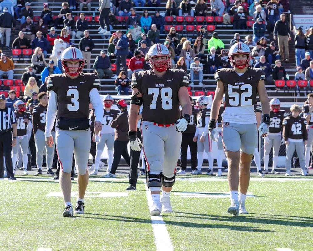 Three senior football players walk side-by-side on the field. The team and crowd mills about on the stands and field in the background.