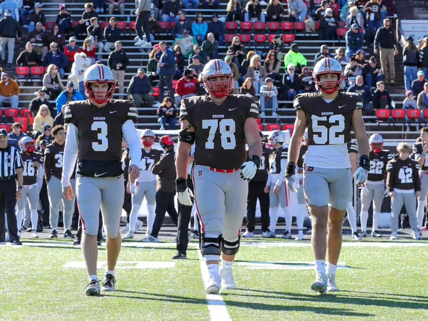 Three senior football players walk side-by-side on the field. The team and crowd mills about on the stands and field in the background.