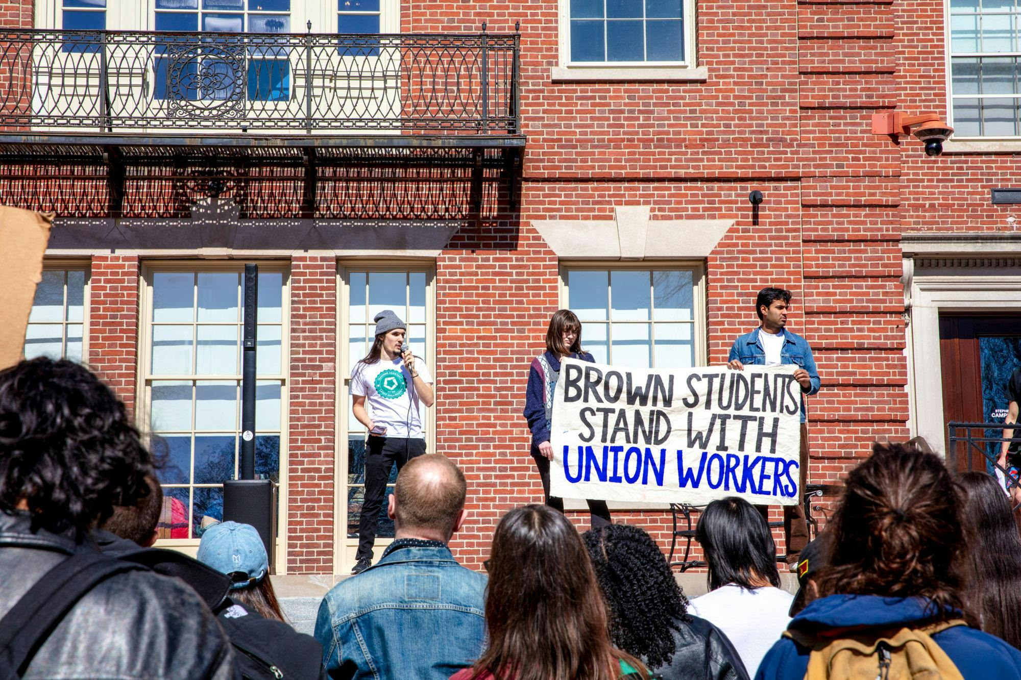On the steps of Campus Center, a speaker and two other people holding up a sign that says "Brown Students Stand with Union Workers."
