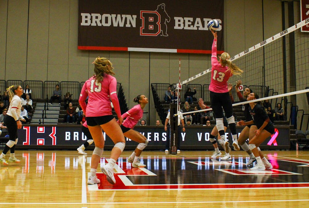 Photo of Brown University women's volleyball players, dressed in pink, at their matchup against Harvard. One player reaches their hand up to the ball as it crosses over the net as the other players watch intently.