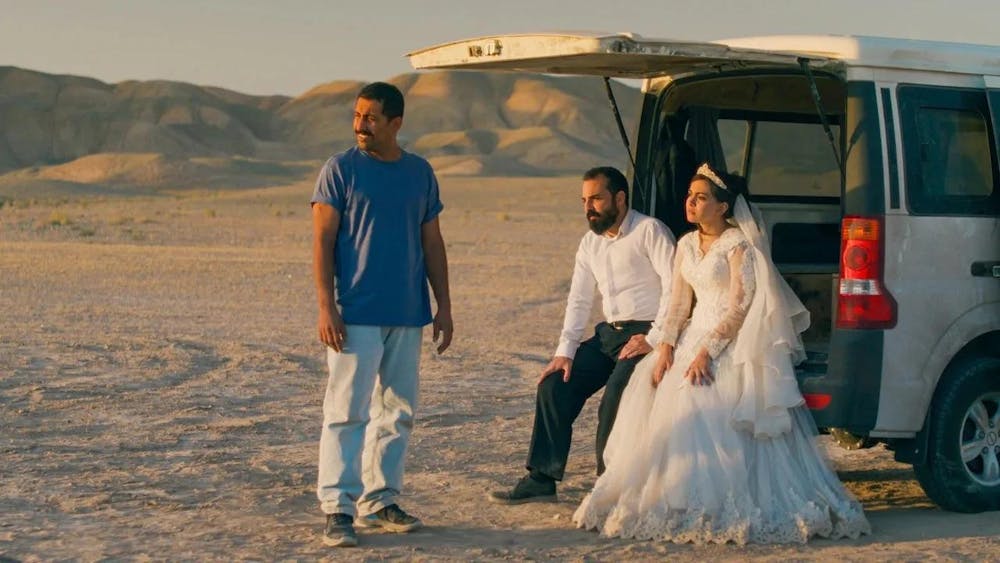 A man in a casual outfit standing in a desert beside a white car with an open trunk where a man in a suit and a woman in a wedding dress are sitting, all looking into the distance.