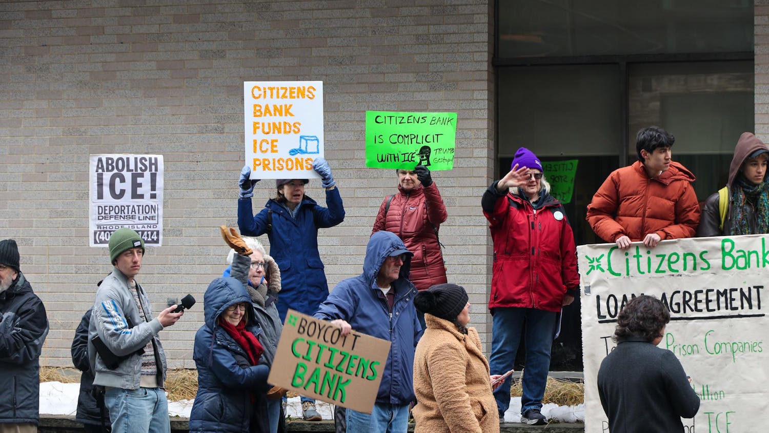 A photo of group of protestors outside of Citizens Bank hold up various signs that read “BOYCOTT CITIZENS BANK,” “CITIZEN BANK FUNDS ICE PRISONS,” “CITIZENS BANK IS COMPLICIT WITH ICE,” and “ABOLISH ICE.”