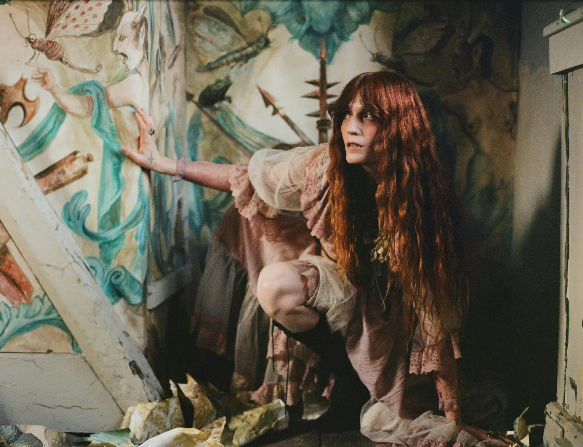 Photo of lead vocalist for Florence and the Machine Florence Welch wearing a dusty pink dress as she crouches and looks to the left in an ornately wallpapered corner.