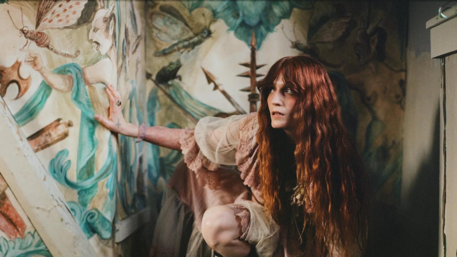 Photo of lead vocalist for Florence and the Machine Florence Welch wearing a dusty pink dress as she crouches and looks to the left in an ornately wallpapered corner.