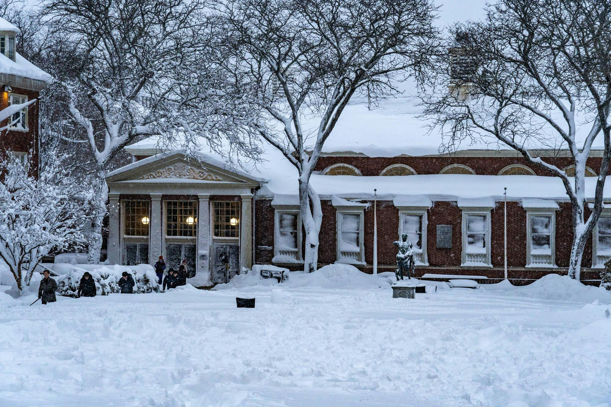 Landscape view of the Sharpe Refectory, the statue of Augustus Caesar and surrounding trees covered in snow with students walking on the left.