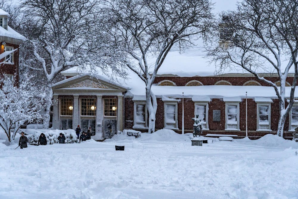 Landscape view of the Sharpe Refectory, the statue of Augustus Caesar and surrounding trees covered in snow with students walking on the left.