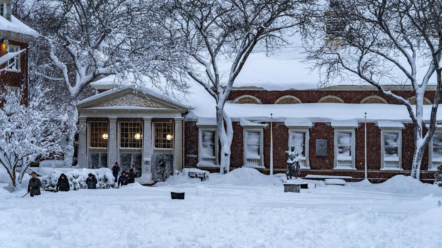 Landscape view of the Sharpe Refectory, the statue of Augustus Caesar and surrounding trees covered in snow with students walking on the left.