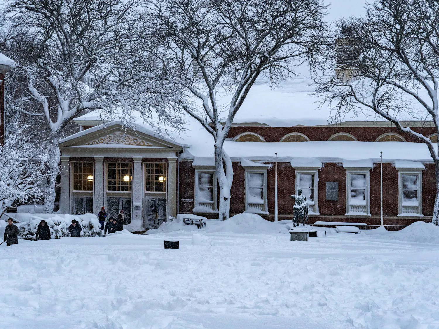 Landscape view of the Sharpe Refectory, the statue of Augustus Caesar and surrounding trees covered in snow with students walking on the left.