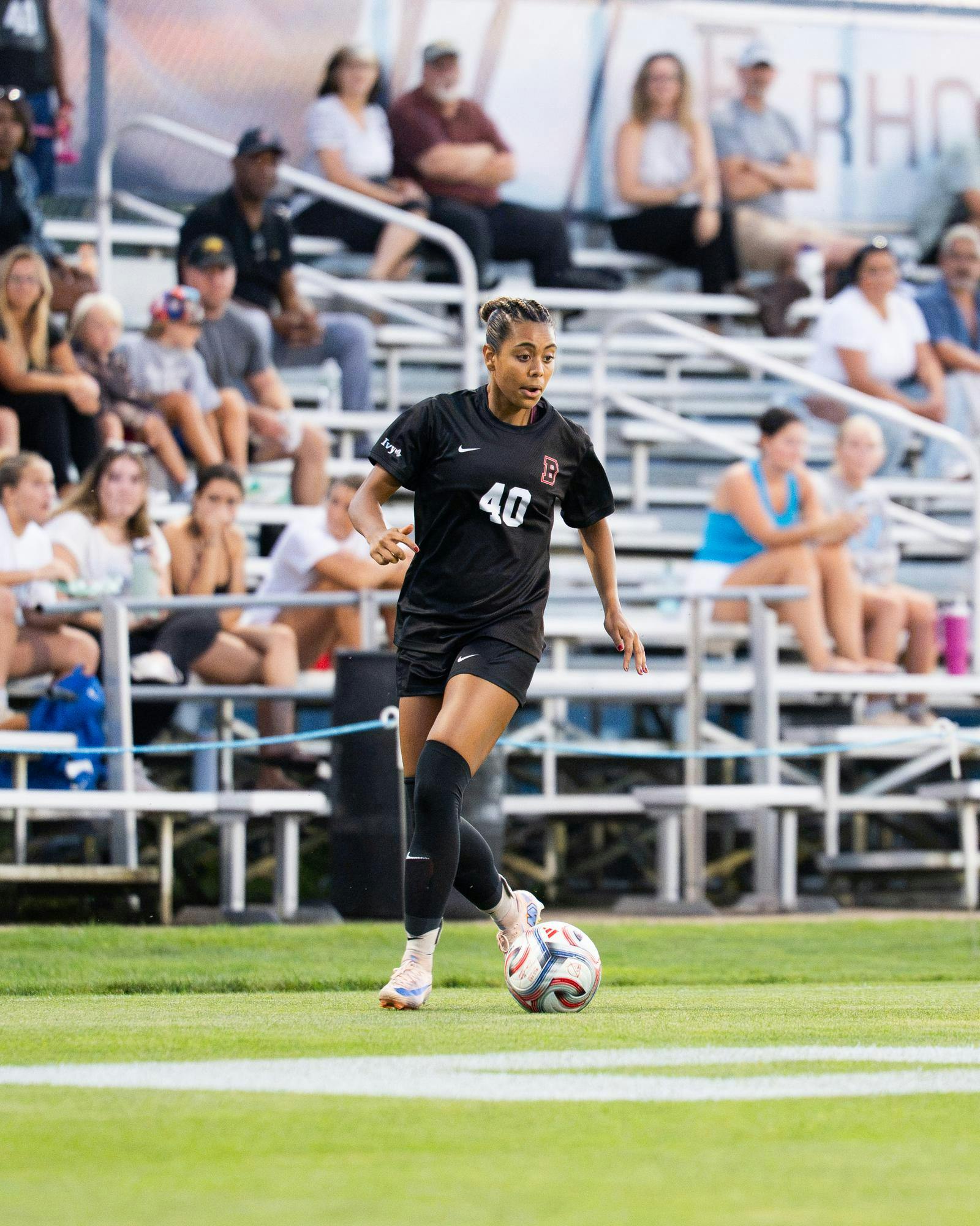 A photograph of a soccer player wearing a Brown jersey on a field. There is a soccer ball at her feet.