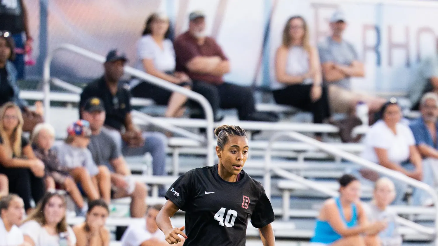 A photograph of a soccer player wearing a Brown jersey on a field. There is a soccer ball at her feet.