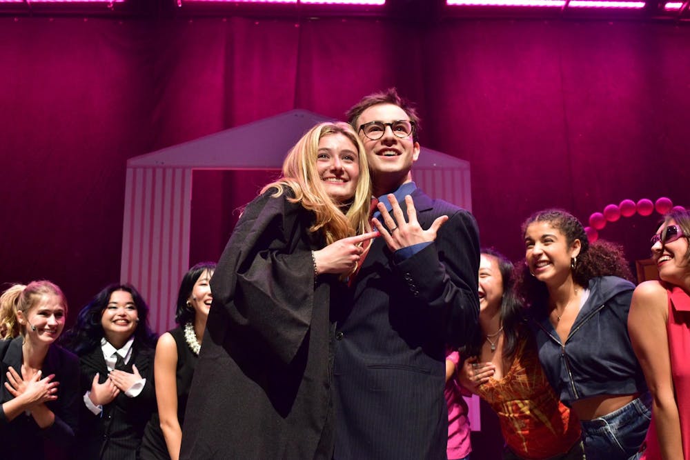 A smiling Elle Woods points at a ring on the finger of her also smiling fiancé, who is wearing a blue striped suit, with a red tie and glasses. 6 other actors surround the pair in a semicircle. 