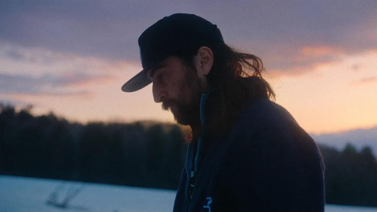 Photo of Noah Kahan wearing a dark blue cap and an unzipped fleece sweater walking in a snowfield with a forest and a warm pink sky in the background. 