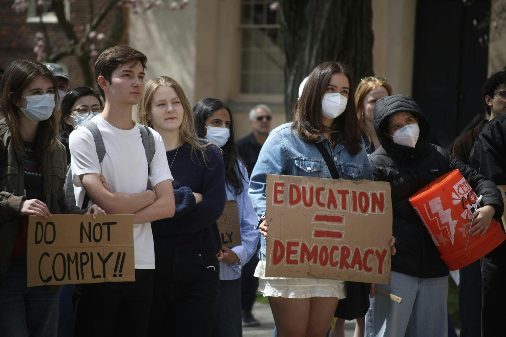 Students stand in protest. One student holds a "Do Not Comply!!" sign, and another masked student holds an "Education = Democracy" sign. 