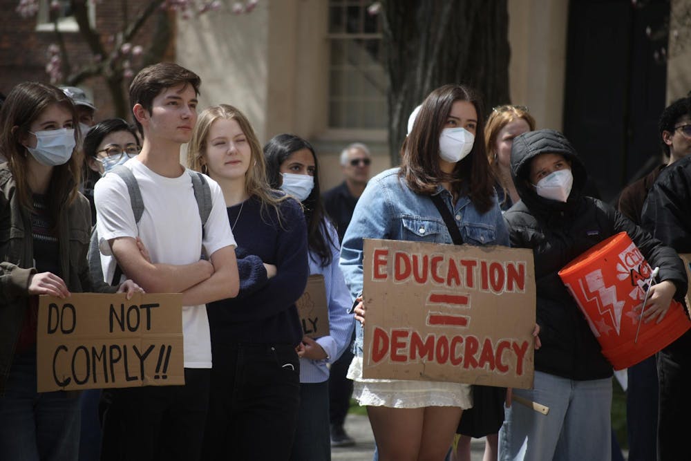Students stand in protest. One student holds a "Do Not Comply!!" sign, and another masked student holds an "Education = Democracy" sign. 
