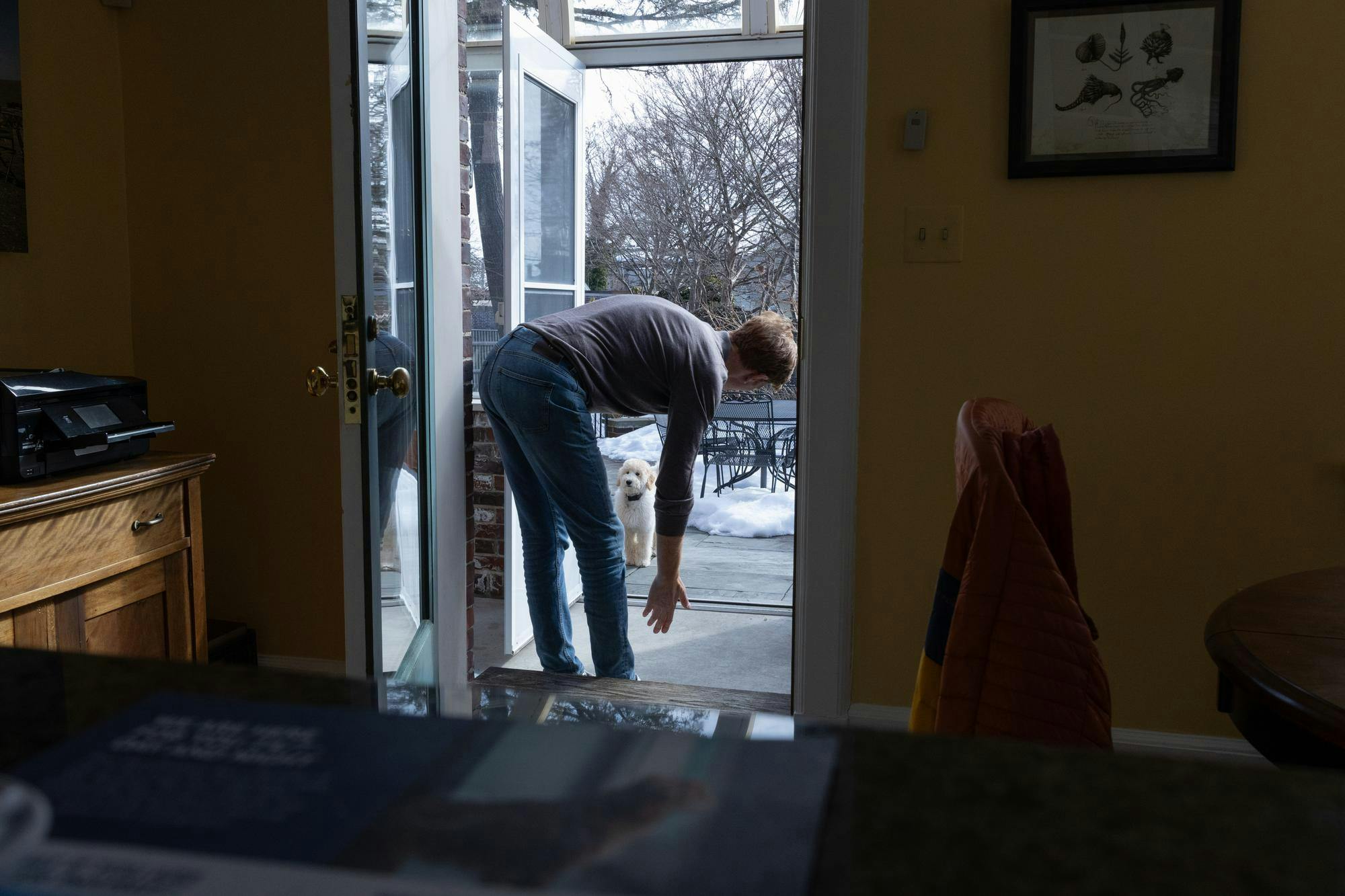 A man standing in a doorframe leans down with his hand stretched out toward a dog.  