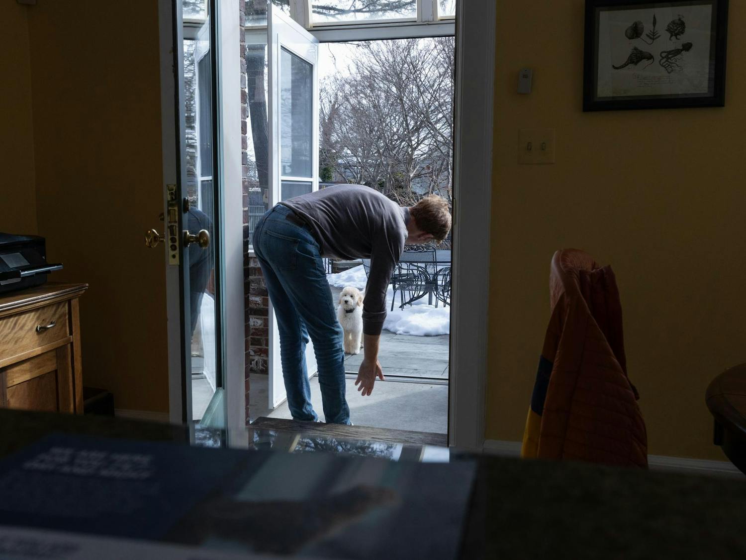 A man standing in a doorframe leans down with his hand stretched out toward a dog.