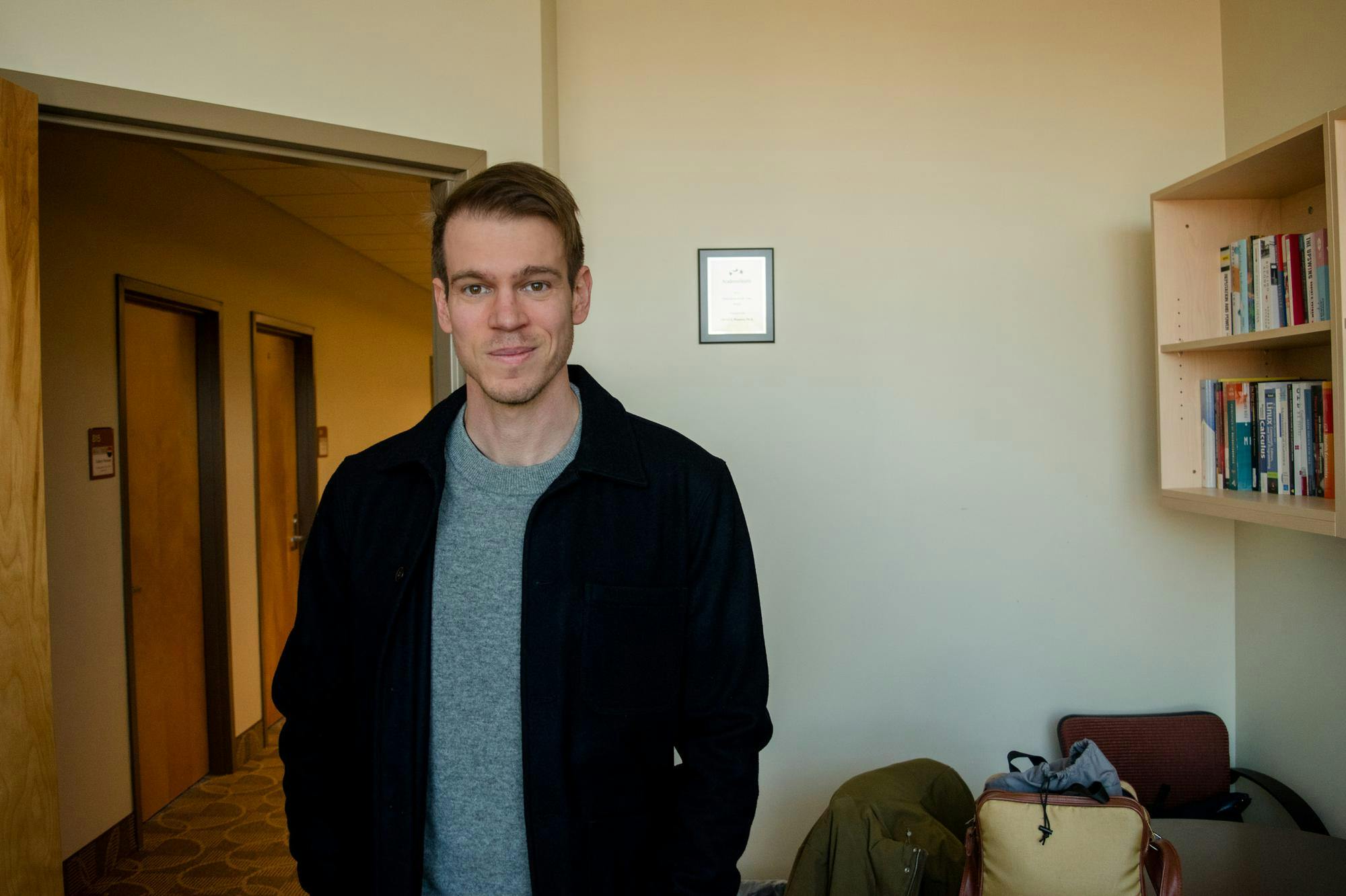 Photo of a man standing in an office with the door open leading to a hallway. The man is wearing a grey shirt and black jacket with his hair swept to the right side and his smile tugs on the right side. There is a bookshelf nailed to the wall on the right filled with books and two bags on a round table at the bottom right and a plaque in the bottom.