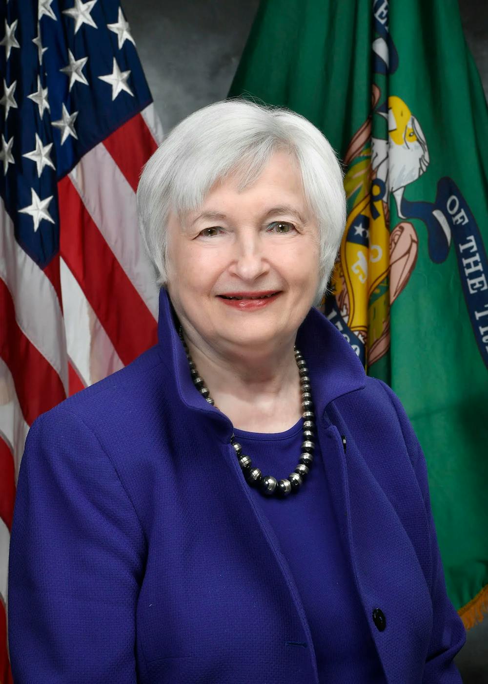 A portrait photo of Janet Yellen who has a gray pixie cut, smiling in front of the American flag. 