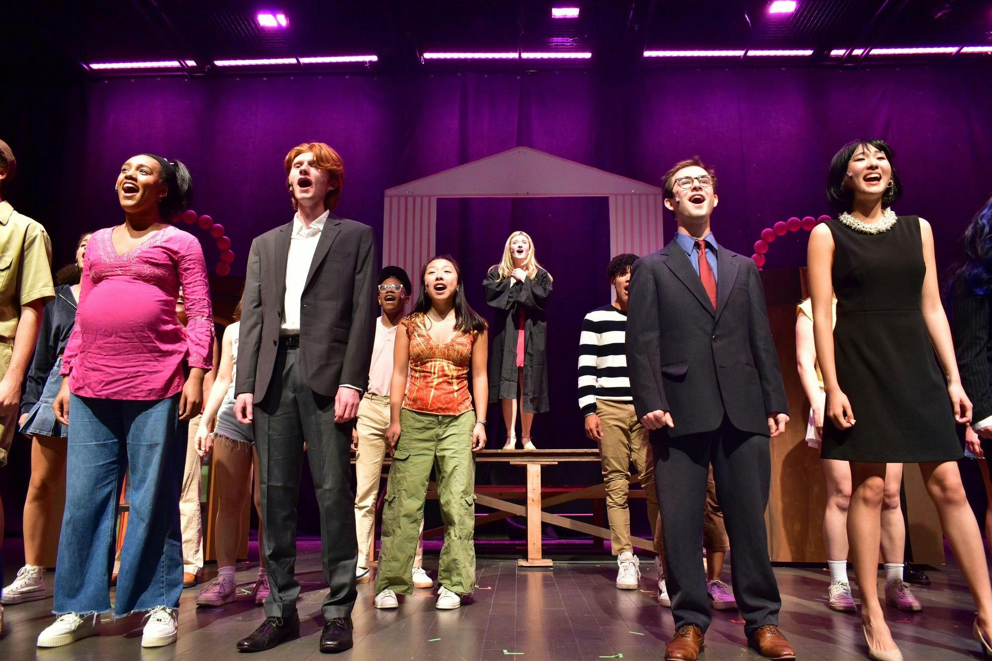 Approximately 14 Legally Blonde actors in a variety of costumes sing facing the audience, with Elle Woods standing in the middle. The stage is lit pink. 