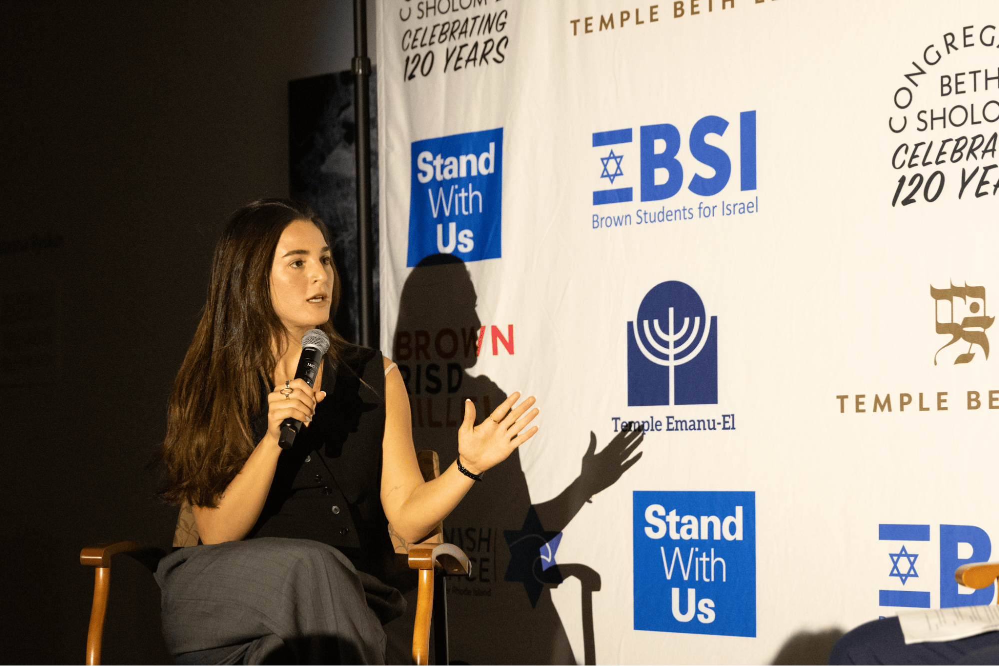 A woman with brown hair and a black top sits on a chair next to a white banner with blue text and logos.