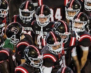 A photograph from an elevated angle of a crowd of Brown football players. White lines on their helmets and shoulders contrast with their brown uniforms.