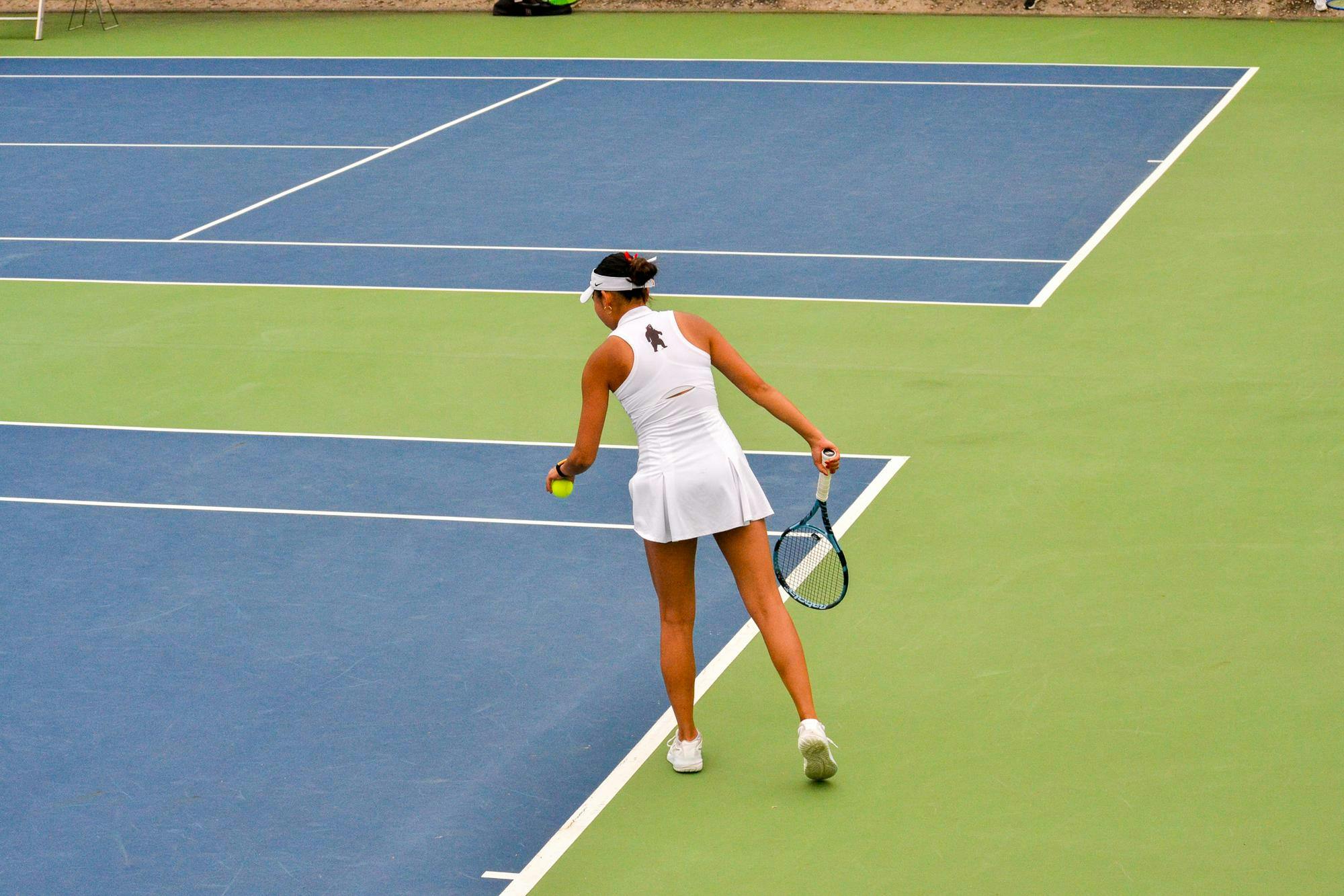 Photo of the back of a woman in a white tank top and white skirt with a brown bear standing up printed on the upper back. She wears a white visor and holds a tennis ball in her left hand and a tennis racket in her right hand, she is holding the tennis ball out about to bounce it.