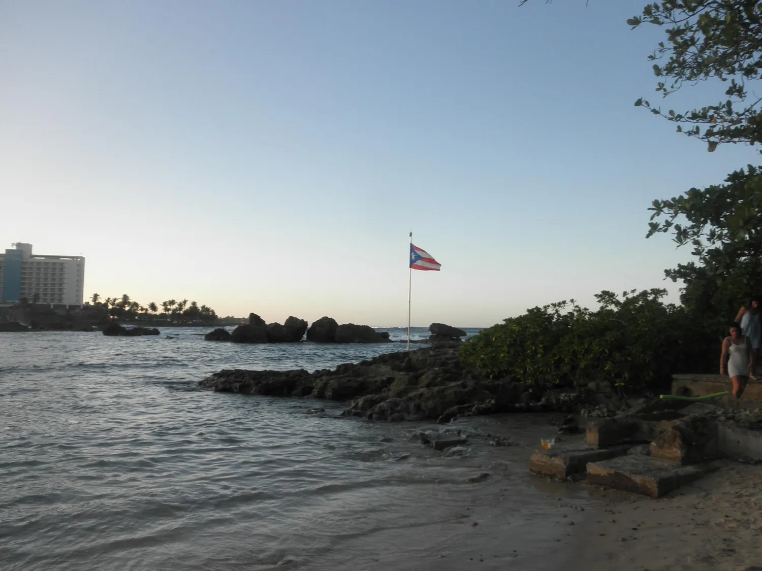 Photo of the beach during sunset with a Puerto Rican flag on a pole on a rock.