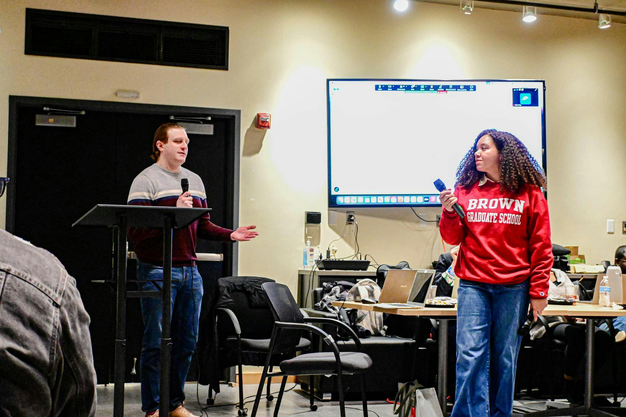 Two Brown University graduate students hold microphones and look at one another.