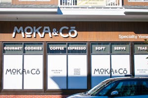 Outside of a store with glass windows covered with white posters with the words "Moka & Co." written across with a QR code in the middle. In the top sections of the windows, the words "Gourmet," "Cookies," "Coffee," "Espresso," "Soft," "Serve," and "Teas" in white bubble letters are posted. On top of it all, on the wooden exterior, the words "Moka & Co." are in large white letters.