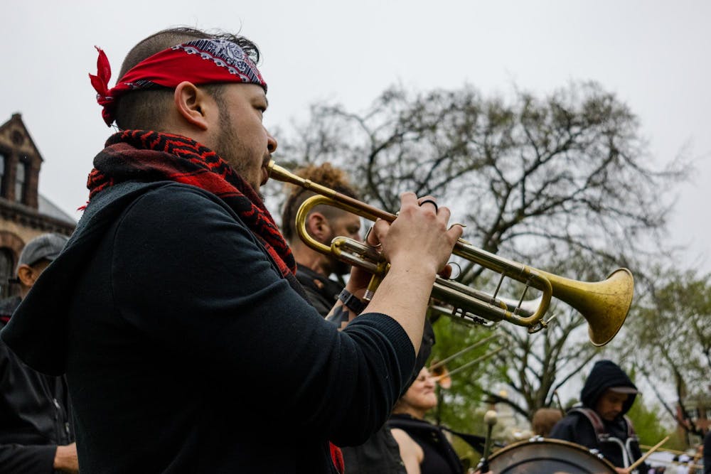 A picture of a person playing a trumpet on the Main Green at Spring Weekend 2025. The person is wearing a black jacket, a red bandana around the neck, and a red bandana tied around the head.