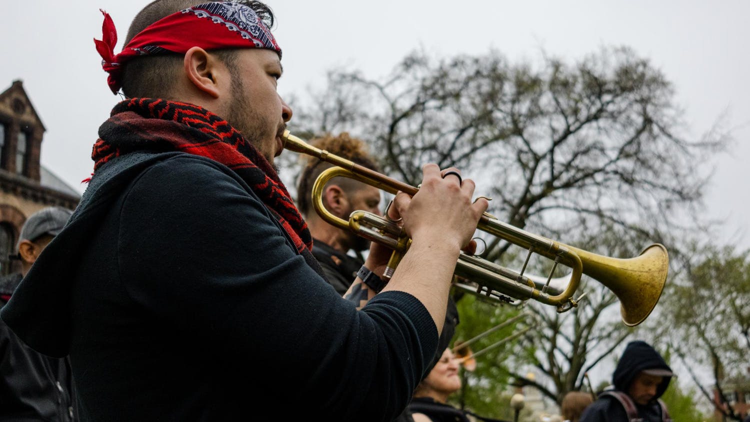 A picture of a person playing a trumpet on the Main Green at Spring Weekend 2025. The person is wearing a black jacket, a red bandana around the neck, and a red bandana tied around the head.