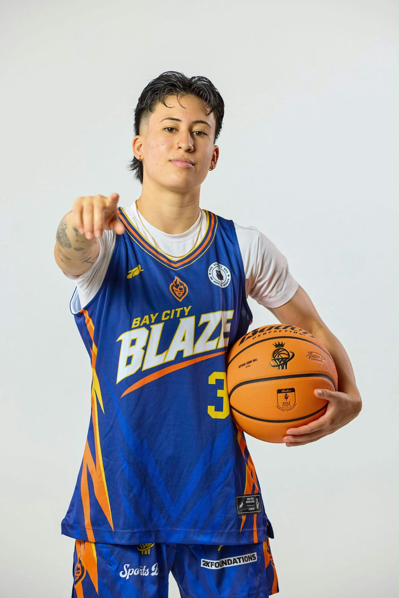 A photo of Isabella Mauricio wearing a blue Bay City Blaze jersey pointing forward and looking into the camera while holding a basketball. 