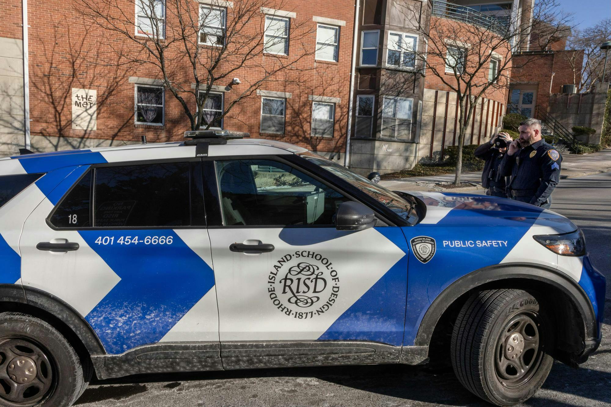 Blue and white Rhode Island School of Design Department of Public Safety car parked upwards on a hill. Two DPS officers stand beside the car. 