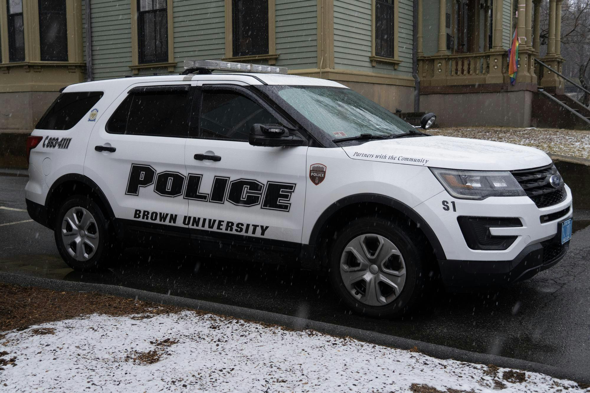 A photo of the side view of a Brown University police car parked on the street while it is snowing outside. 