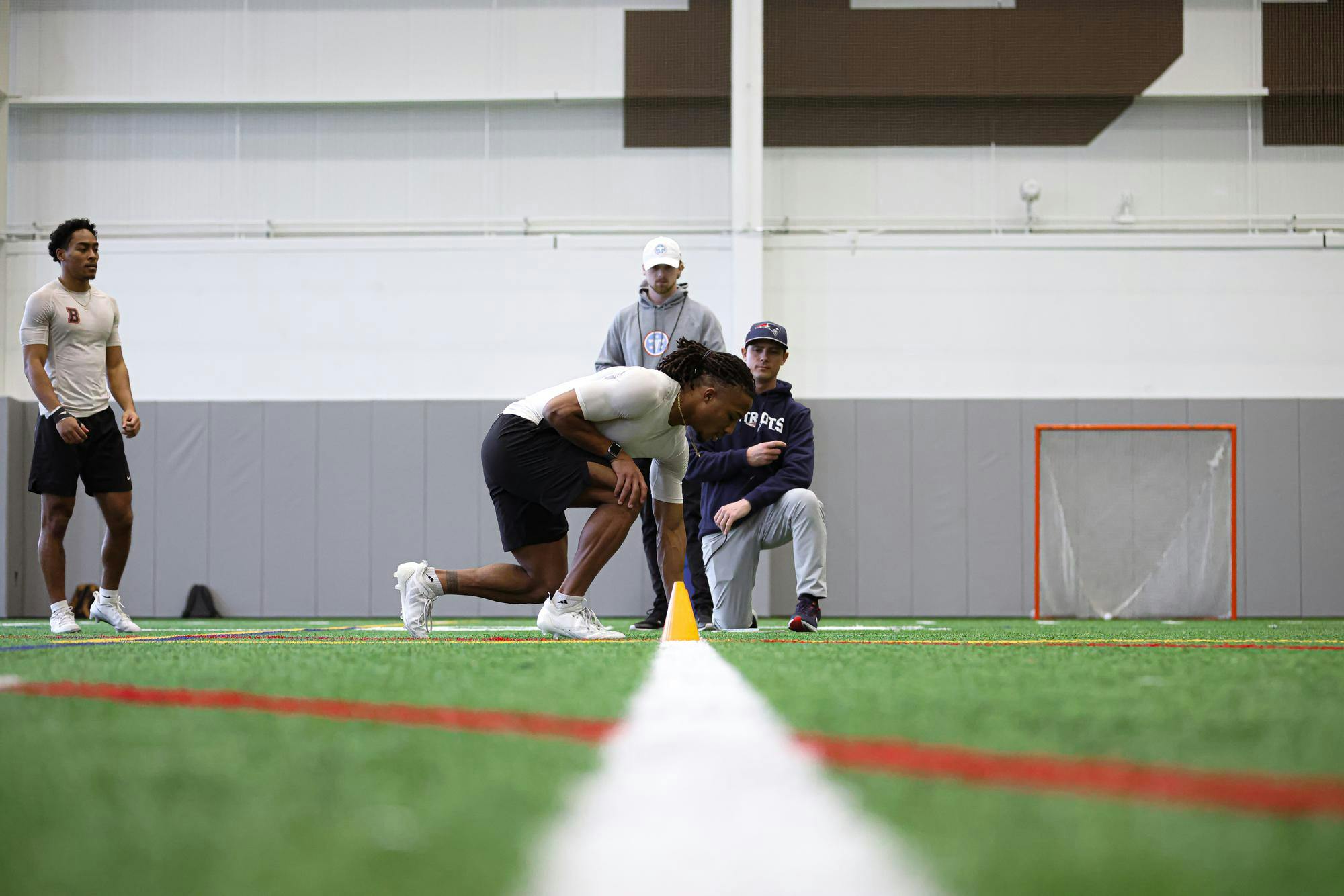A man kneels at a start line on a turf field in front of two football scouts.